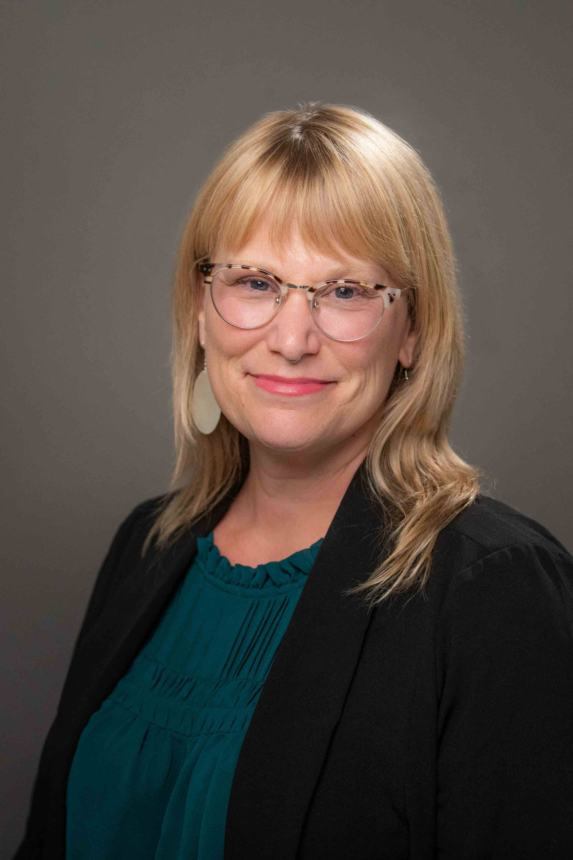 Holly wears a teal blouse, black blazer, and has tortoiseshell glasses